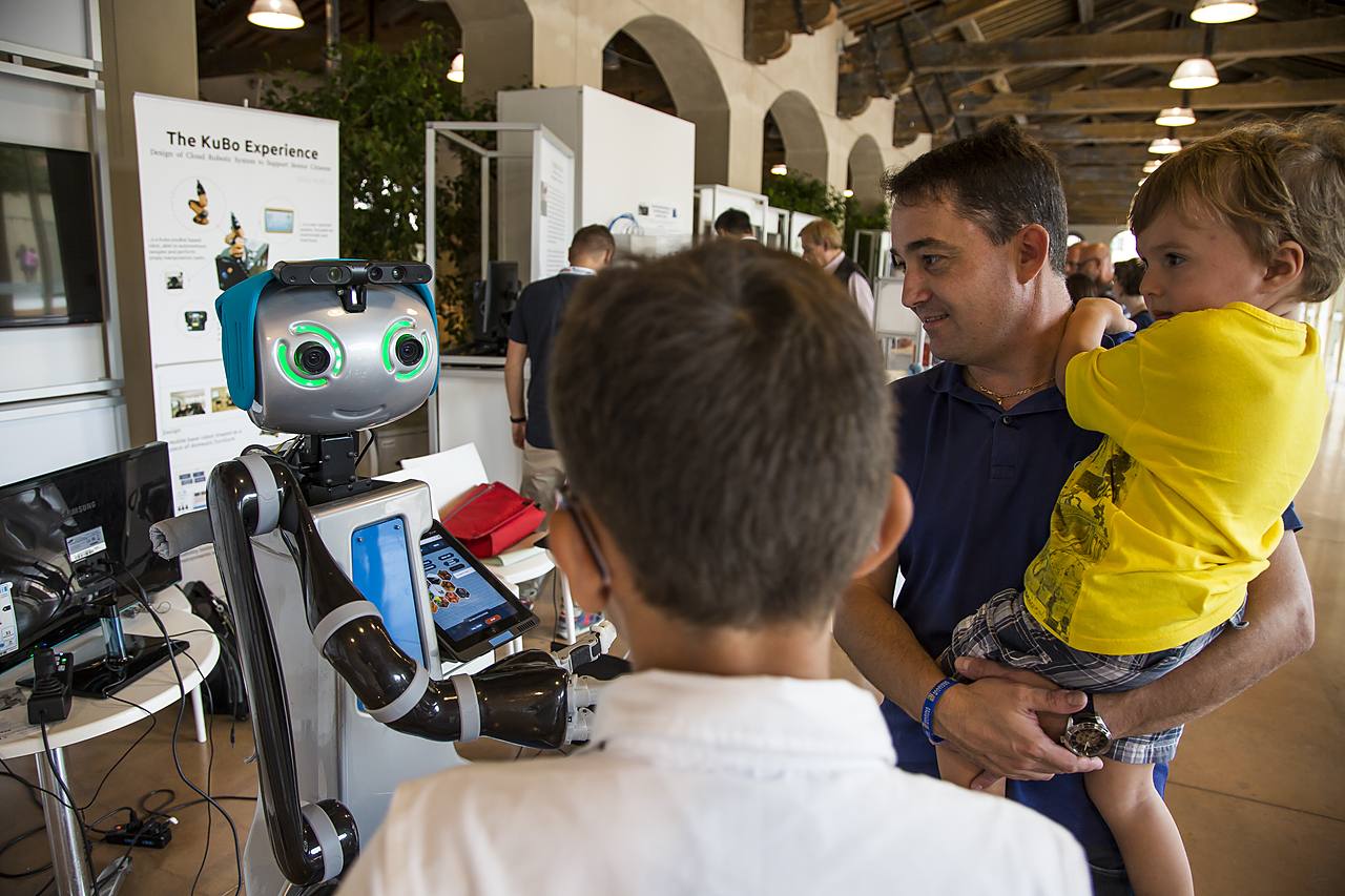 Un padre e un bambino osservano un piccolo robot in uno stand del Festival della Robotica: tecnologie e robotica assistiva per la disabilità.