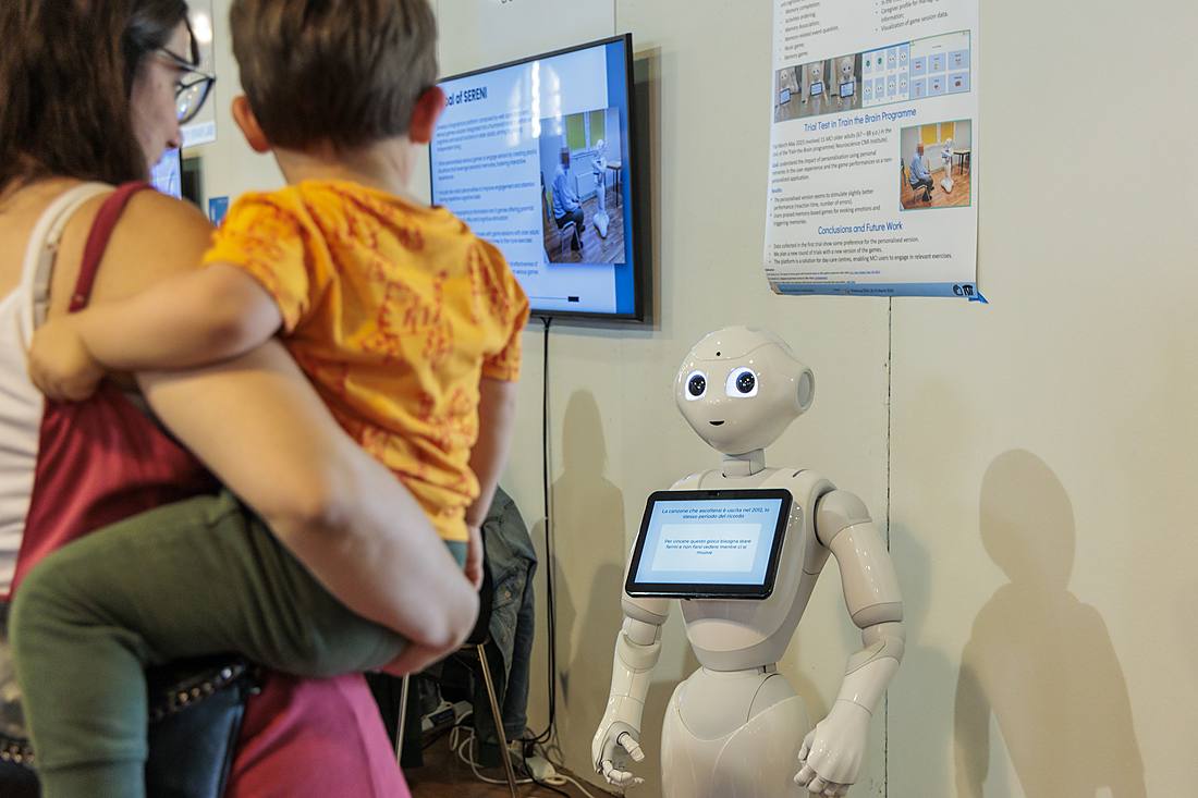 A mother holding a child looks at the Pepper humanoid robot at the Festival della Robotica: assistive robotics and technologies for disability.