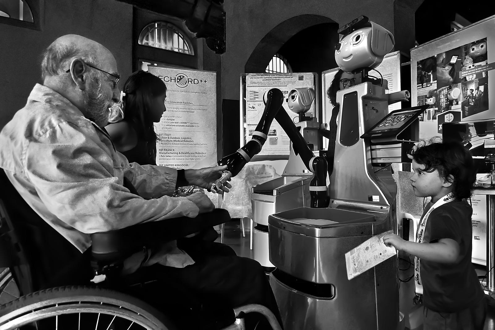 A service robot arm helps an elderly person in a wheelchair while a little girl looks on: assistive robotics for everyday independence.